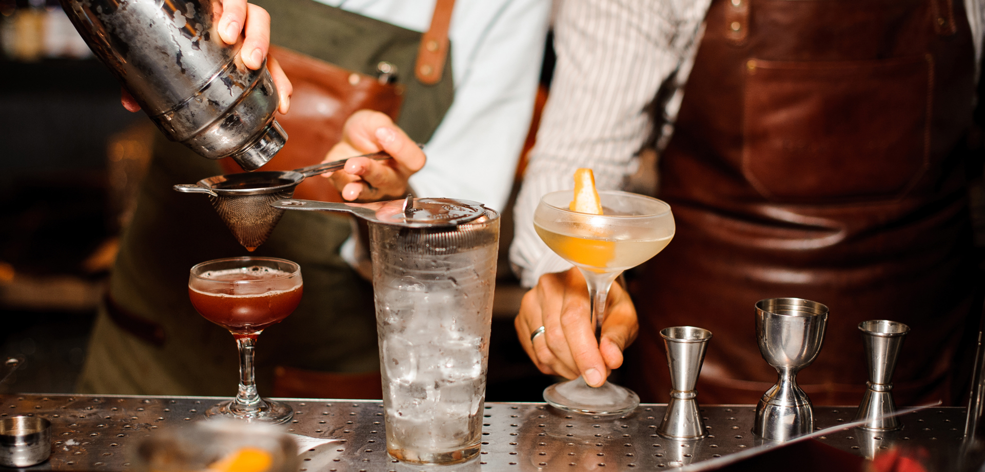 Two bartenders are stood behind a bar, wearing aprons. They are making cocktails using equipment such as a cocktail strainer. 