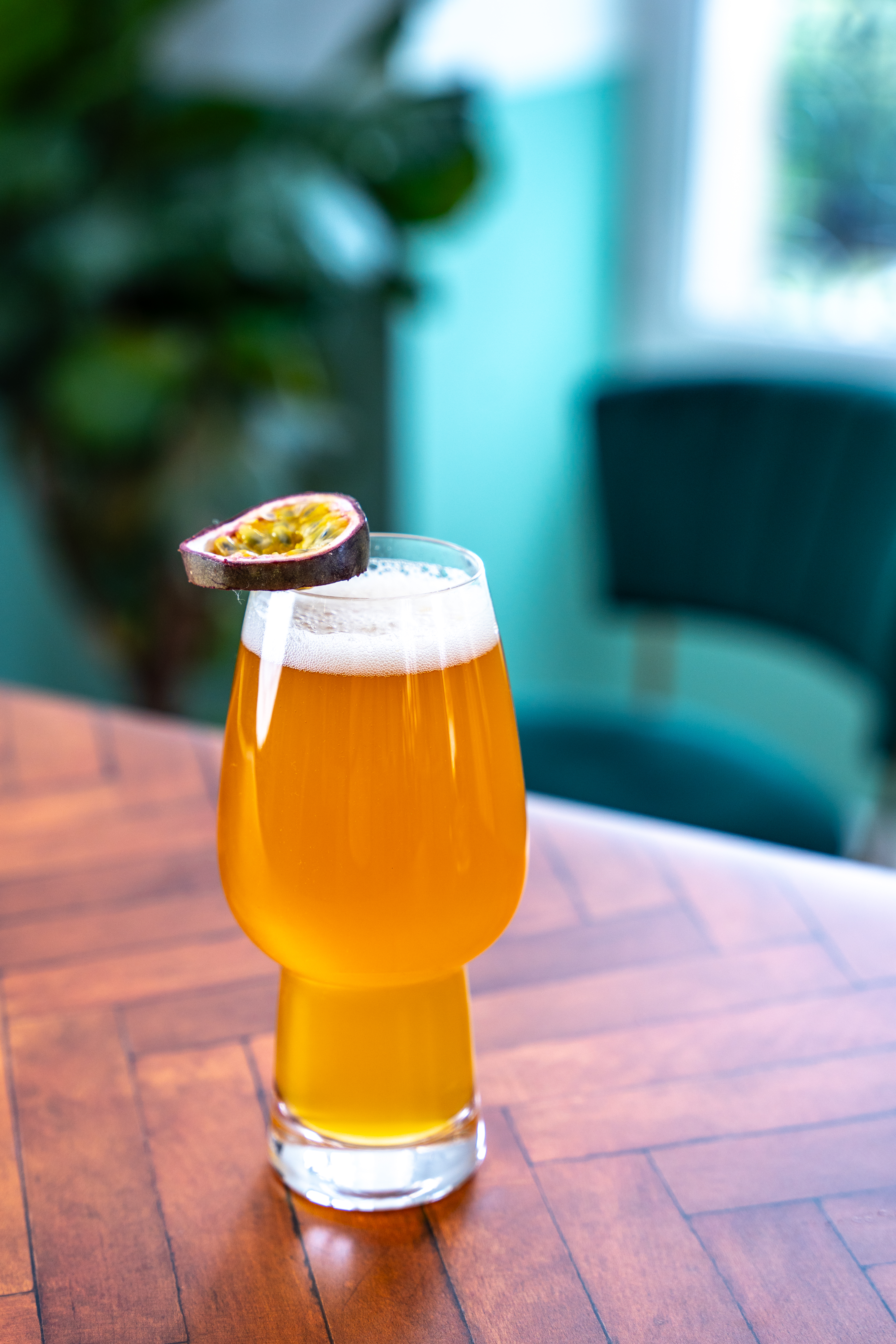A beer glass sits on a polished wooden surface. The drink is topped with a passionfruit slice. In the background there is a chair and a potted plant. 
