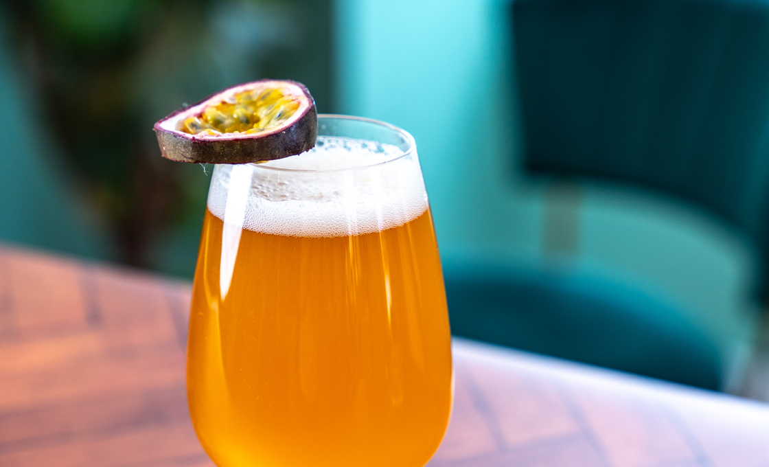 A beer glass sits on a polished wooden surface. The drink is topped with a passionfruit slice. In the background there is a chair and a potted plant. 