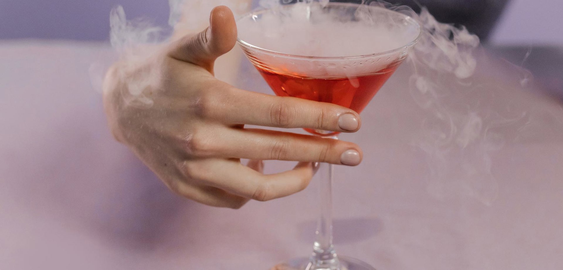 A female hand wrapped around a martini cocktail glass. The glass is filled with red liqued and is smoking with dry ice.