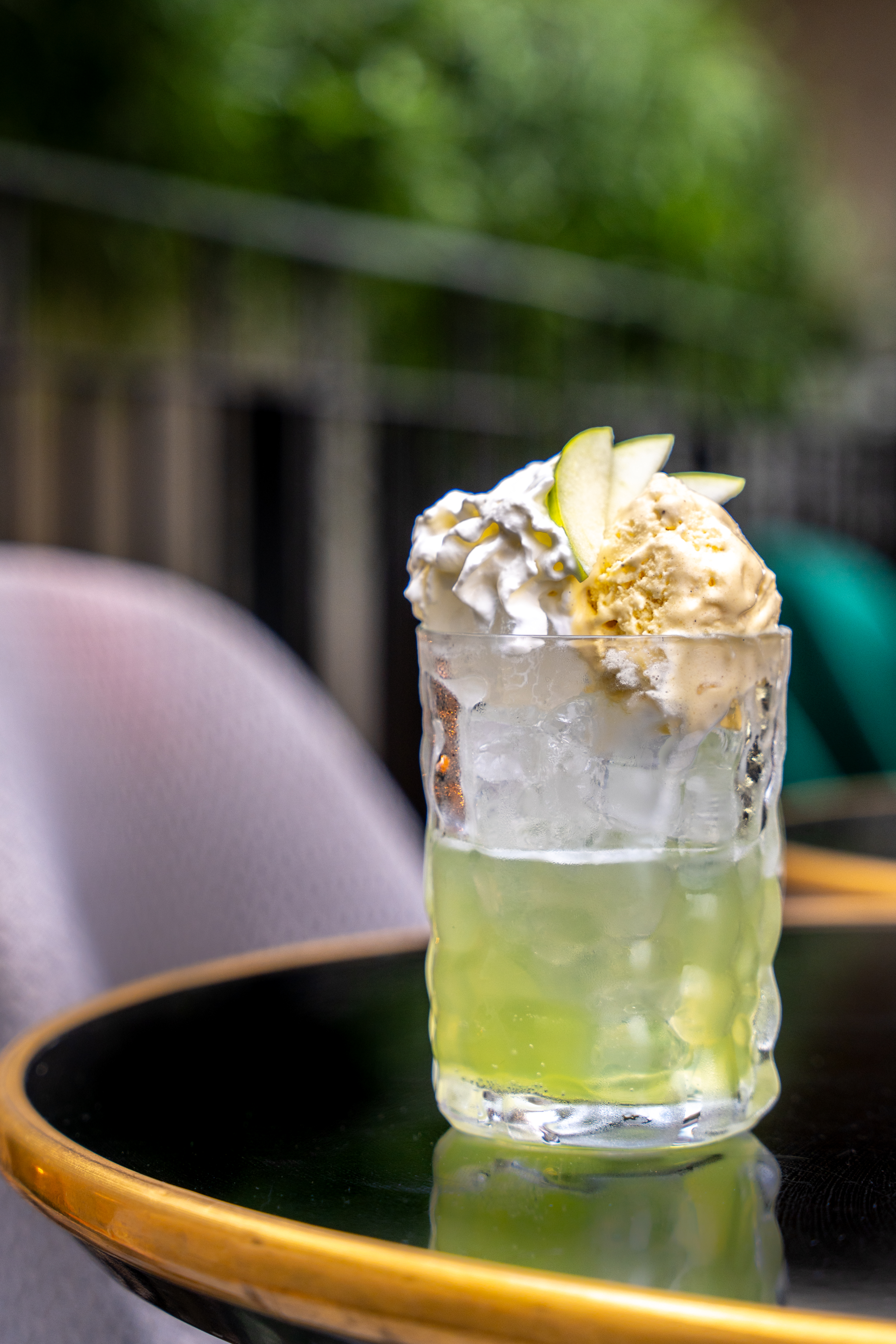 On the edge of a black coffee table there is a clear glass that contains a green drink. The drink is topped with whipped cream. In the background there are bar seats. 