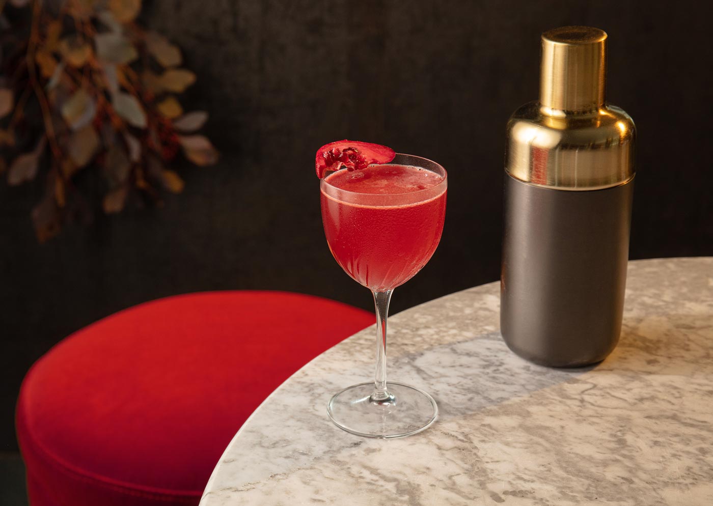 A short coupe glass sits on the edge of a marble table, placed next to a cocktail shaker. The glass contains  a red drink topped with a pomegranate slice. A red bar stool is placed until the table, and a plant can be seen in the background.