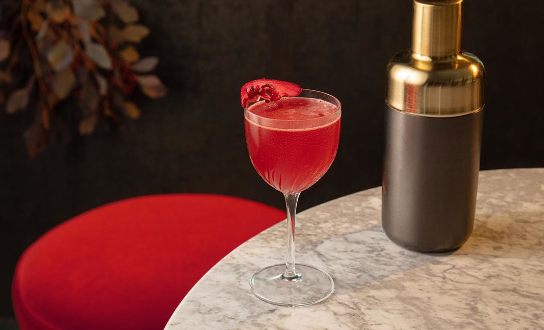 A short coupe glass sits on the edge of a marble table, placed next to a cocktail shaker. The glass contains  a red drink topped with a pomegranate slice. A red bar stool is placed until the table, and a plant can be seen in the background.