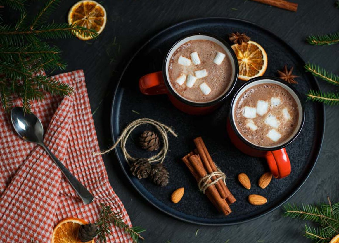 Two mugs of hot chocolate, garnished with marshmallows sitting on a black tray. The tray is decorated with dried orange, fir tree brances and cinnamon sticks.