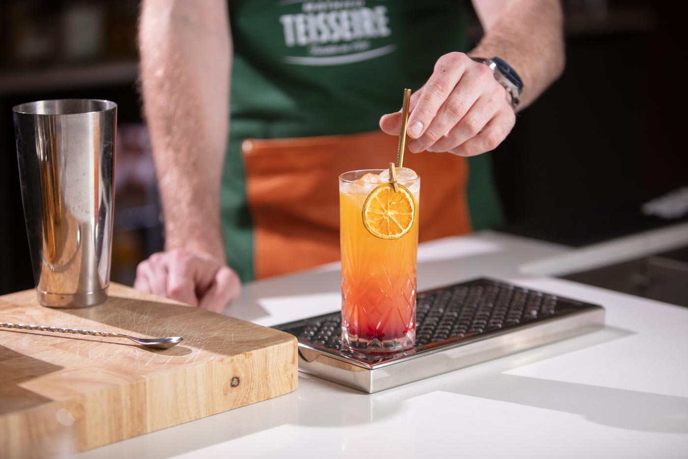 On top of a bar grate is a highball glass. The glass contains an orange drink. On the edge of the glass is an orange slice held in place by a gold peg. A bartender is placing a gold straw into the glass.