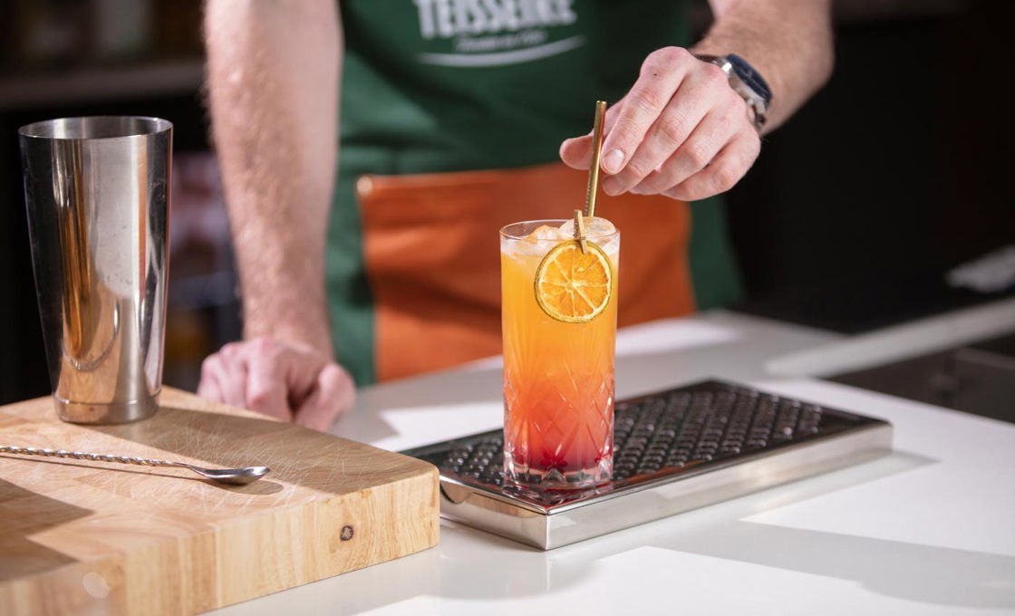 On top of a bar grate is a highball glass. The glass contains an orange drink. On the edge of the glass is an orange slice held in place by a gold peg. A bartender is placing a gold straw into the glass.
