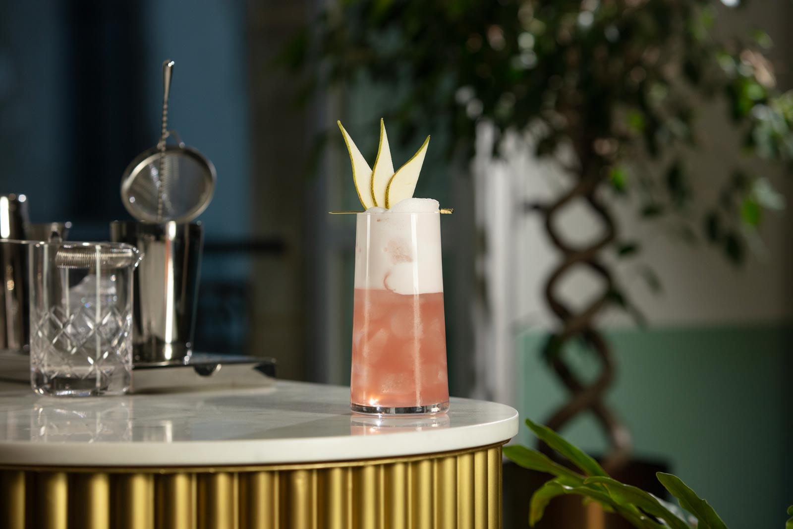 A tall glass is on the edge of a bar. The glass contains a pink drink topped with white foam and a pear fan on a gold pick. Also on the bar we can see various pieces of cocktail making equipment.