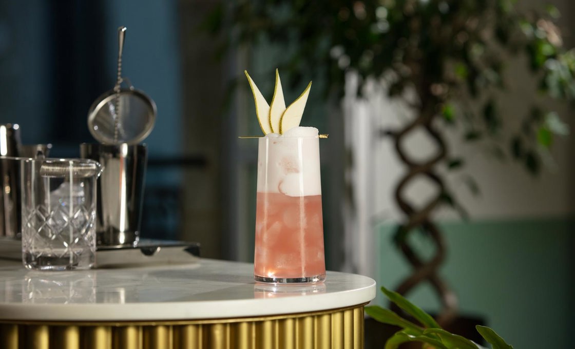 A tall glass is on the edge of a bar. The glass contains a pink drink topped with white foam and a pear fan on a gold pick. Also on the bar we can see various pieces of cocktail making equipment.