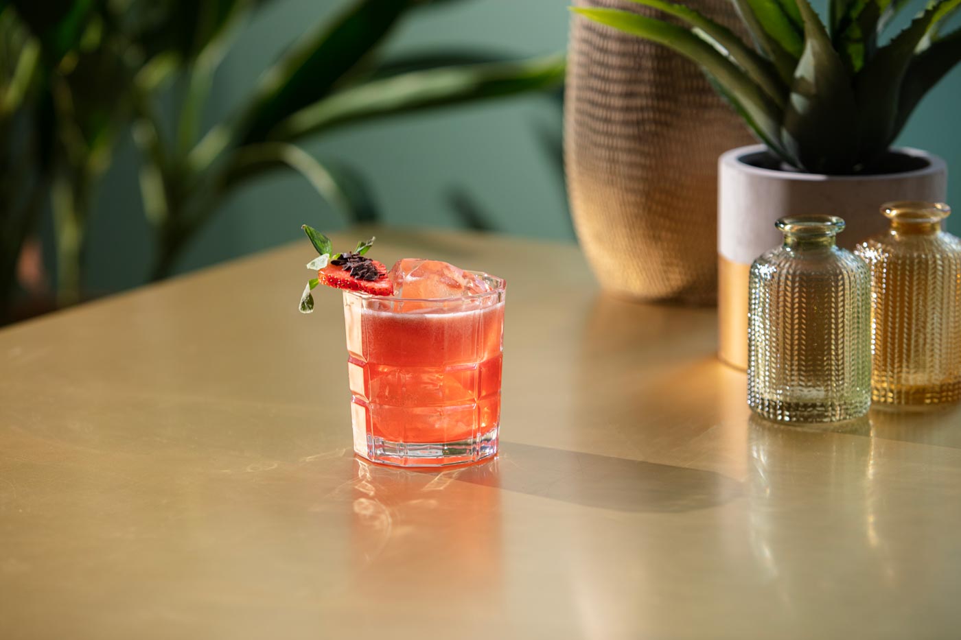 A short rocks glass is on a gold table. The glass contains a pink drink that has been topped with a slice of strawberry and some chocolate shavings. In the background we can see some plant pots.