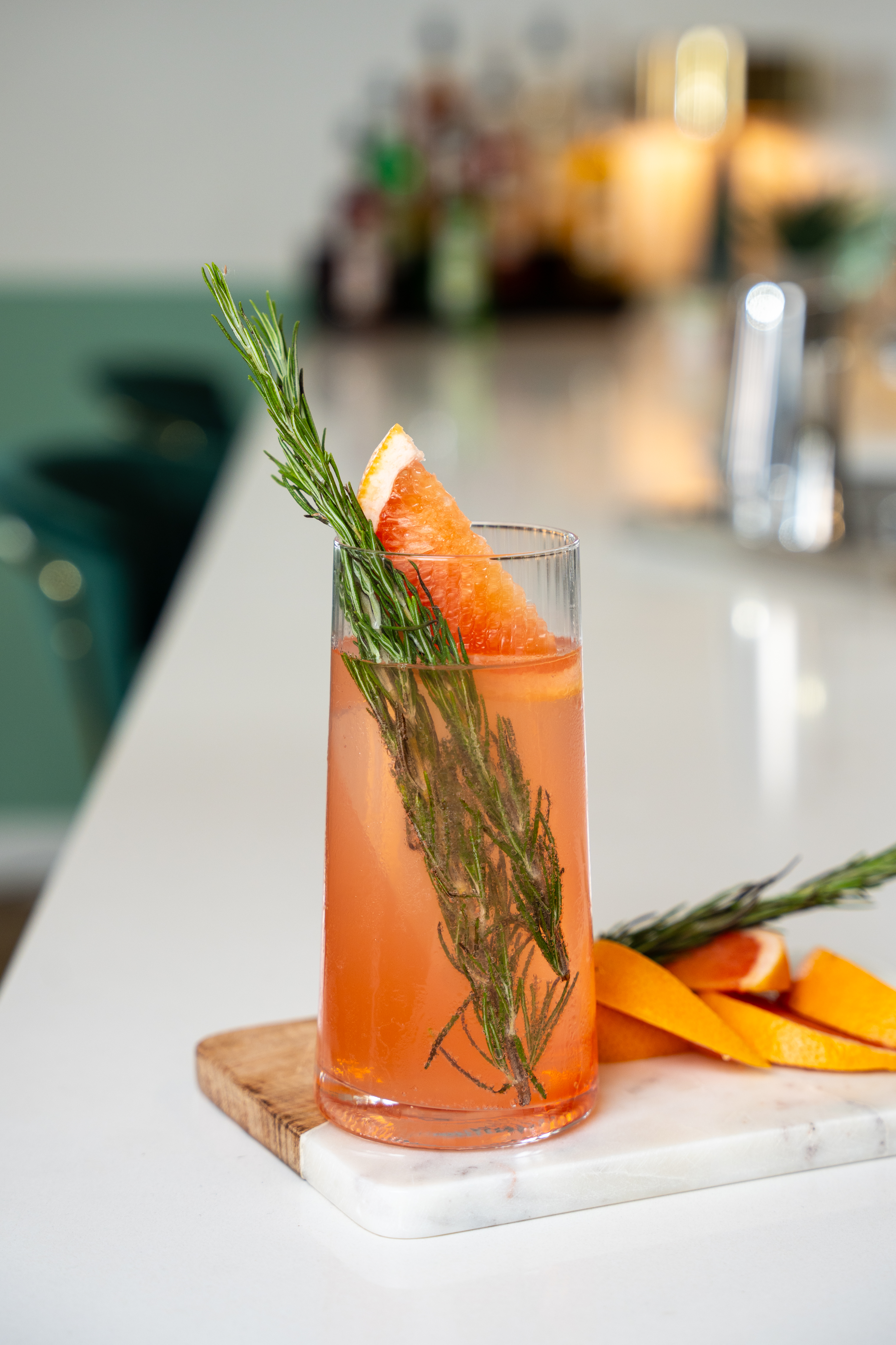A tumbler glass is placed atop a serving tray on a long marble bar surface. Inside the glass is a pink coloured drink, rosemary and a grapefruit wedge.