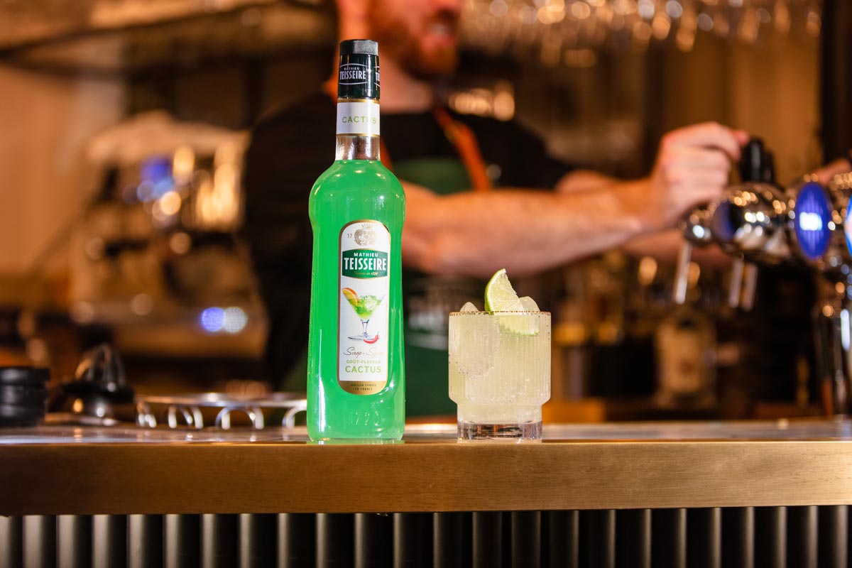 On a bar sits a short glass containing a white drink, topped with a wedge of lime. Next to the glass is a bottle of Mathieu Teisseire cactus syrup. In the background there are bar taps, with a bartender stood behind wearing a green apron.