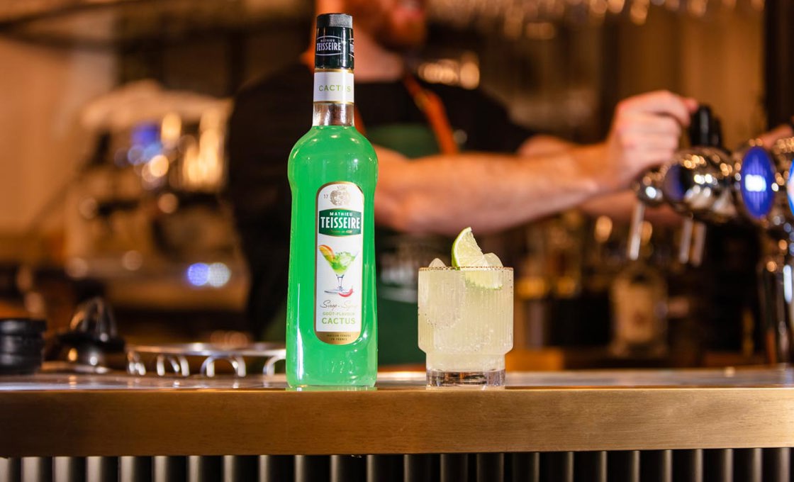 On a bar sits a short glass containing a white drink, topped with a wedge of lime. Next to the glass is a bottle of Mathieu Teisseire cactus syrup. In the background there are bar taps, with a bartender stood behind wearing a green apron.