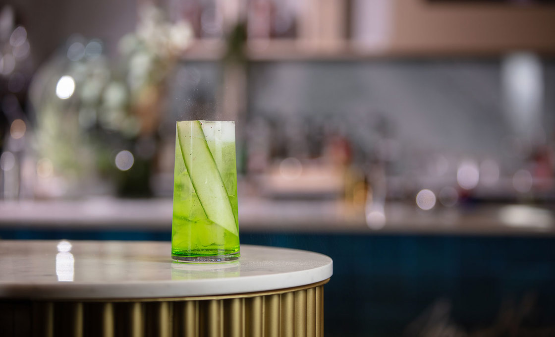 A tall glass is on the edge of a bar and contains a green drink. Inside the drink is a slice of cucumber. In the background we can see a wall witrh shelves that contain Mathieu Teisseire syrups.