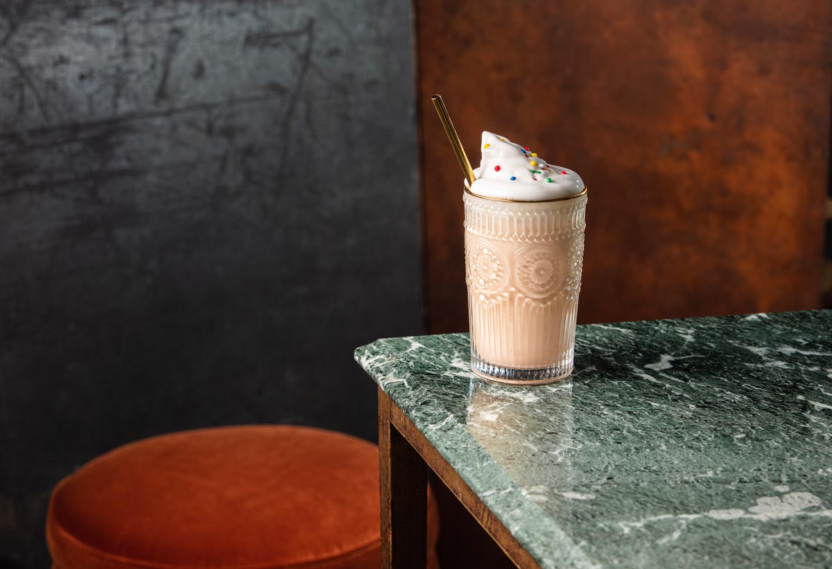 On the edge of a green marble table a milkshake glass has been placed. The glass contains a milkshake and is topped with whipped cream. There is a gold metal straw in the drink. In the background there is an orange bar stool.
