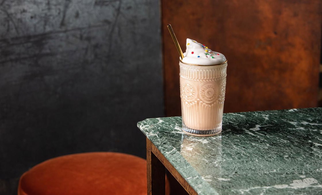 On the edge of a green marble table a milkshake glass has been placed. The glass contains a milkshake and is topped with whipped cream. There is a gold metal straw in the drink. In the background there is an orange bar stool.