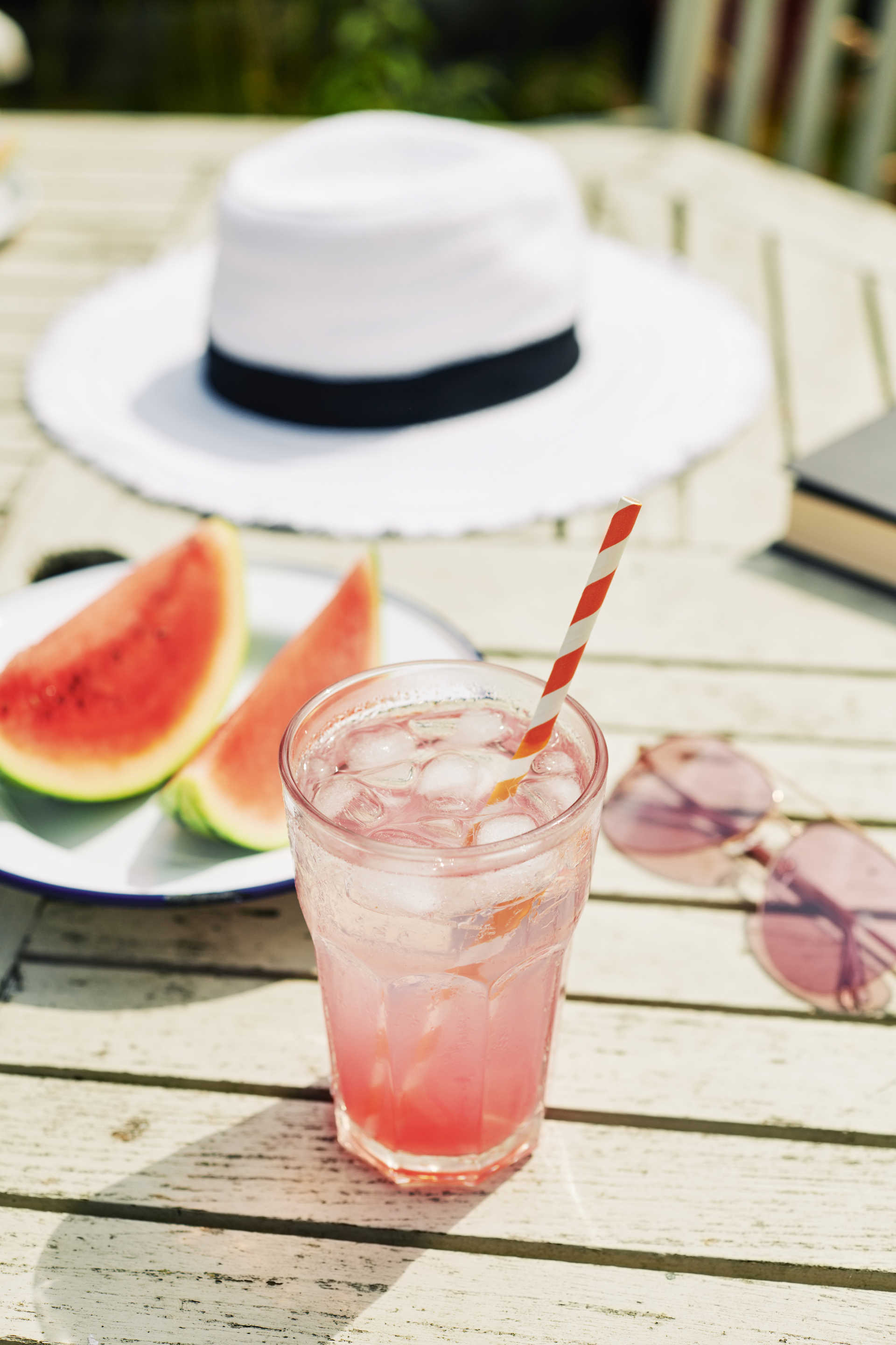 A glass of a melonium cocktail on a table outdoors along with a sliced watermelon, sunglasses and a hat