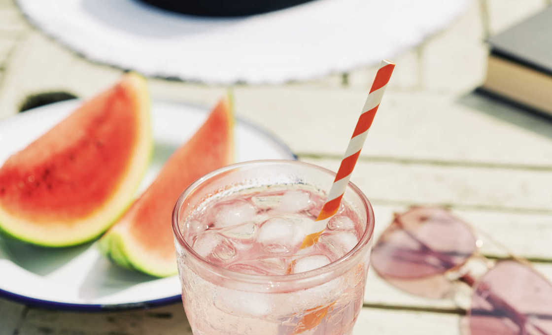 A glass of a melonium cocktail on a table outdoors along with a sliced watermelon, sunglasses and a hat
