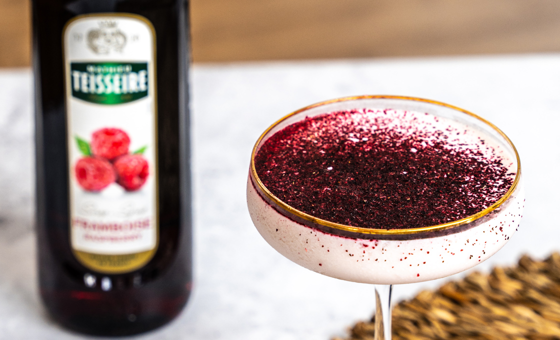 A martini glass is on a marble surface, inside the glass is a pink coloured drink topped with raspberry powder. Next to the glass there is a bottle of Mathieu Teisseire raspberry syrup. 