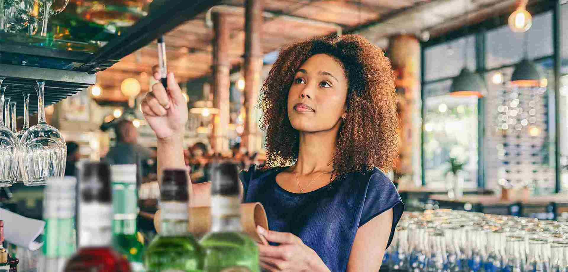 A woman stands behind a bar holding a clipboard and pen. She holds the pen in the air and looks up, as though counting stock.