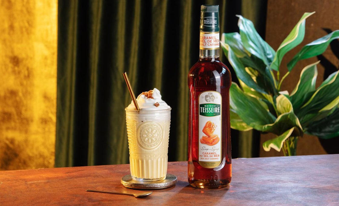 A milkshake glass containing a milkshake drink topped with whipped cream and a gold metal straw is placed on a silver coaster. In front of the glass is a long stirring spoon. Next to the glass is a bottle of Mathieu Tiesseire caramel syrup.