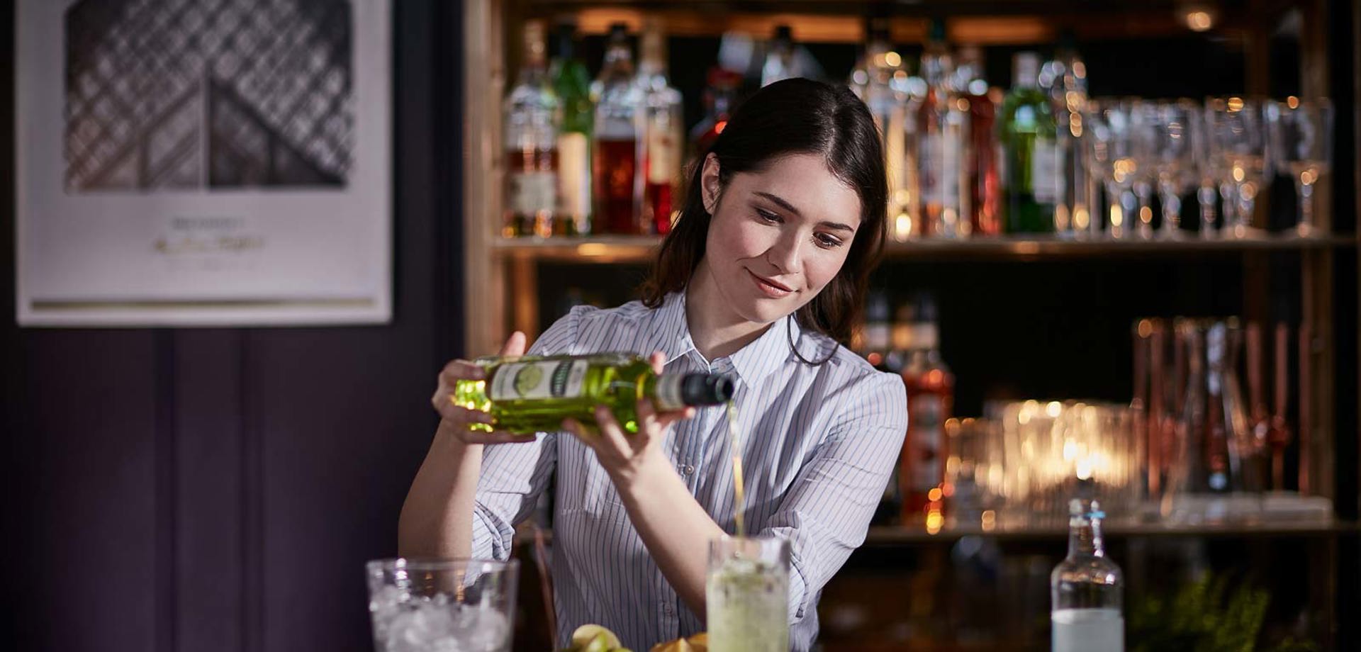 A female bartender standing behing a bar, pouring cocktail syrup into a glass. On the bar there is a glass filled with ice and bowls of lemon and lime wedges.