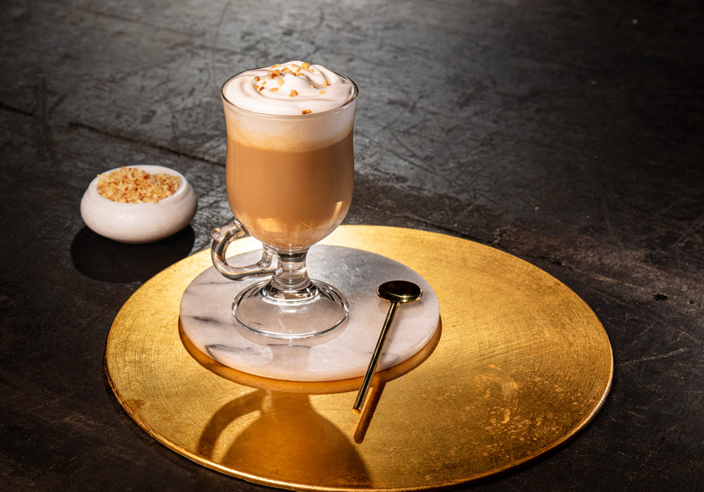 A latte glass contains coffee and is topped with whipped cream. It is sitting on a marble coaster. Placed next to the glass is a large gold stirring spoon. In the background is a small pot containing the drinks garnish.