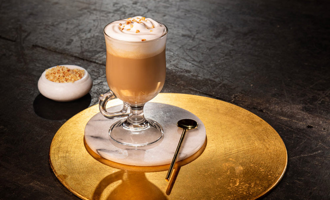 A latte glass contains coffee and is topped with whipped cream. It is sitting on a marble coaster. Placed next to the glass is a large gold stirring spoon. In the background is a small pot containing the drinks garnish.