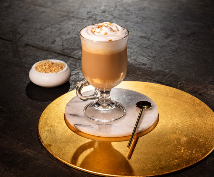 A latte glass contains coffee and is topped with whipped cream. It is sitting on a marble coaster. Placed next to the glass is a large gold stirring spoon. In the background is a small pot containing the drinks garnish.