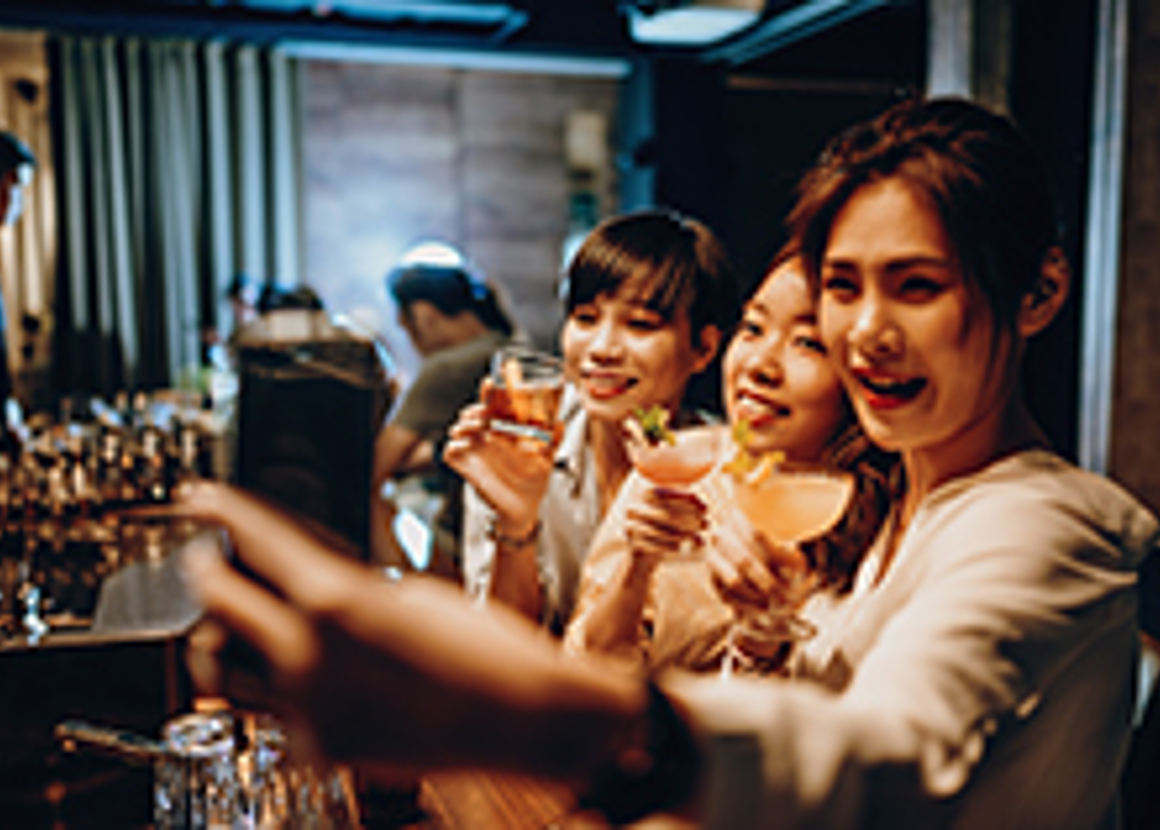 A group of 3 women are holding cocktails in a cocktail bar and taking a selfie.