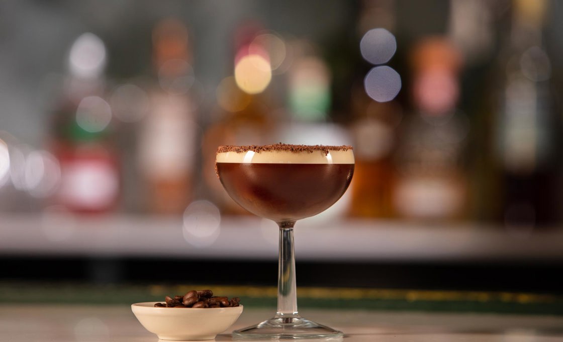 A short coupe glass is placed on a bar. The bar contains a chocolate coloured drink and is topped with chocolate shavings.