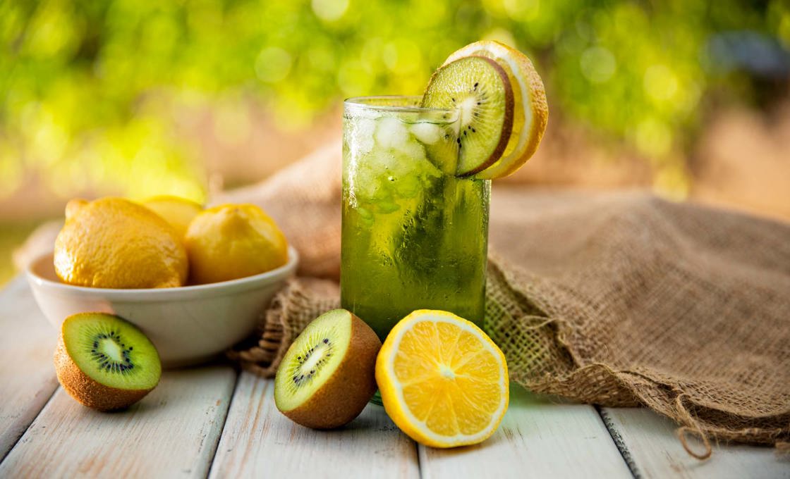 A glass of kiwi lime lemonade with ice on a table with lemons, limes and kiwis