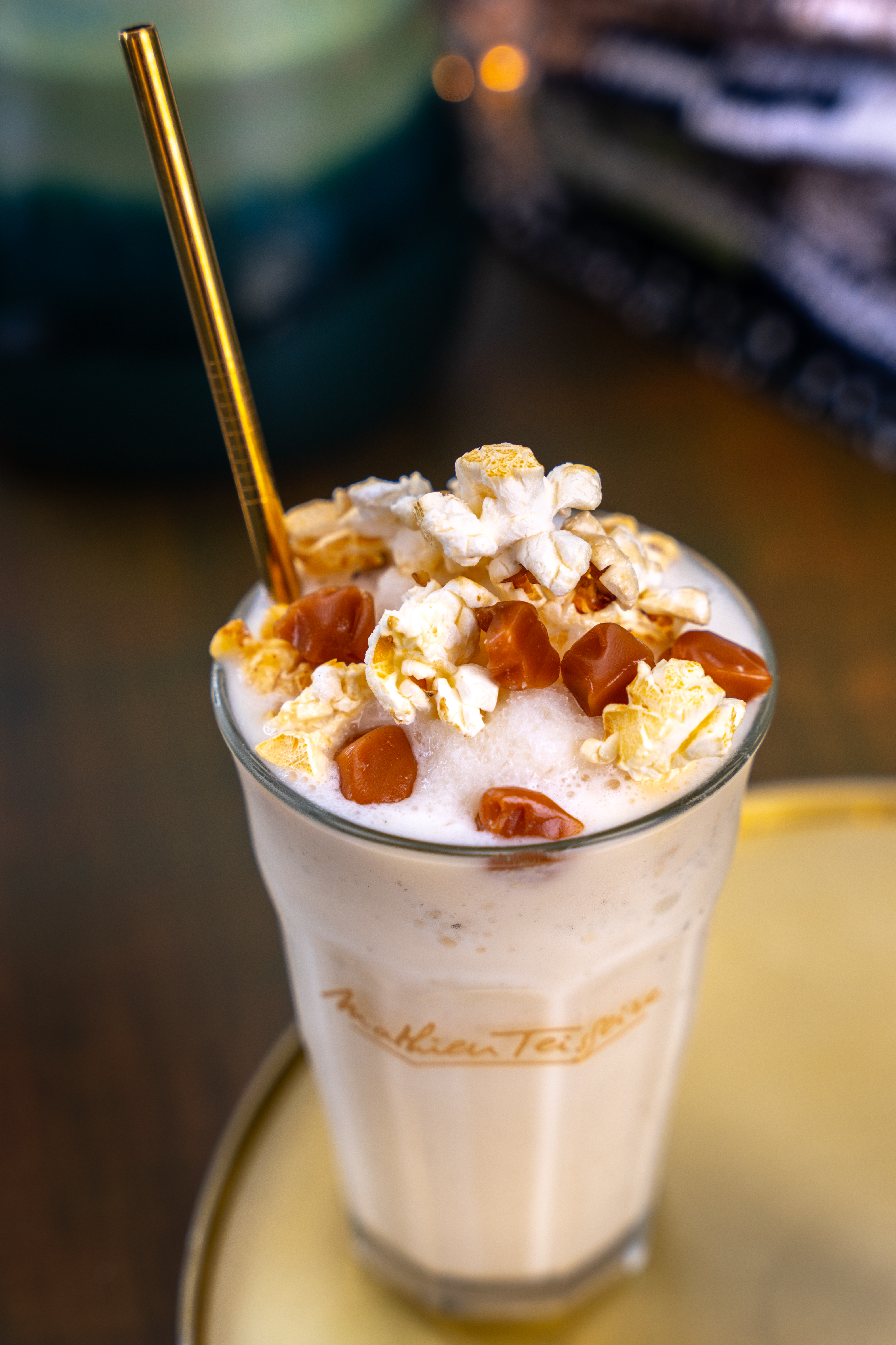 On the edge of a gold tray there is a Mathieu Teisseire glass, containing a white coloured milkshake. The milkshake is topped with cream, popcorn and chunks of caramel.