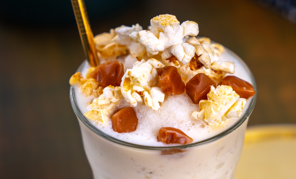 On the edge of a gold tray there is a Mathieu Teisseire glass, containing a white coloured milkshake. The milkshake is topped with cream, popcorn and chunks of caramel.