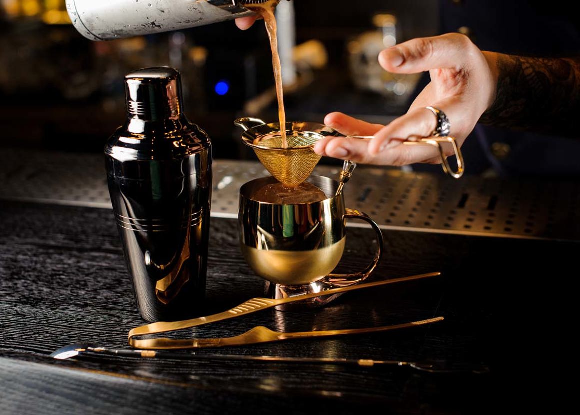 On a dark surface, a drink is being strained into a mug. On the surface there is also a cocktail shaker and other drink making equipment.