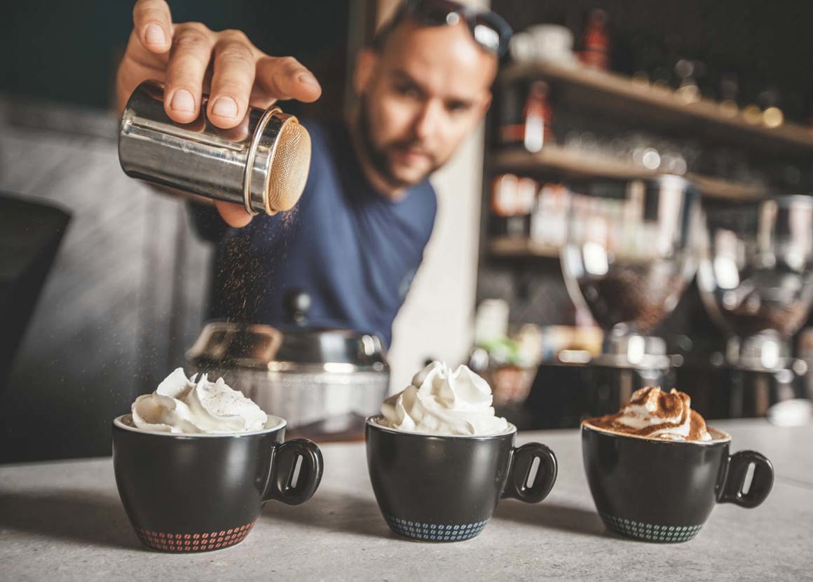 A male barista standing behind a counter shaking chocolate powder over three hot chocolates that he has made. The hot chocolates are in black cups and are garnished with whipped cream.