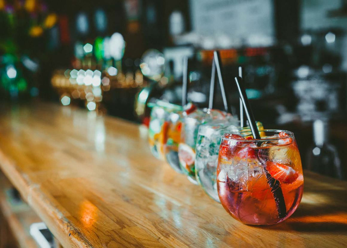 Along the length of a wooden bar there is a row of short glasses. The glasses all contain fizzy drinks of different colours.