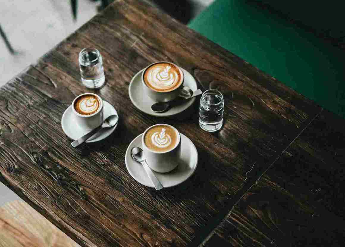 Three cups of coffee are placed in the centre of a wooden table. In the background there is a green velvet chair and a tall plant.