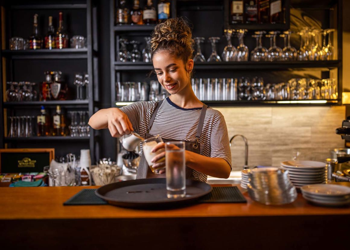 A female barista standing behind a counter. She is pouring milk into a highball glass. There is a black tray in front of her with a glass of water on it.
