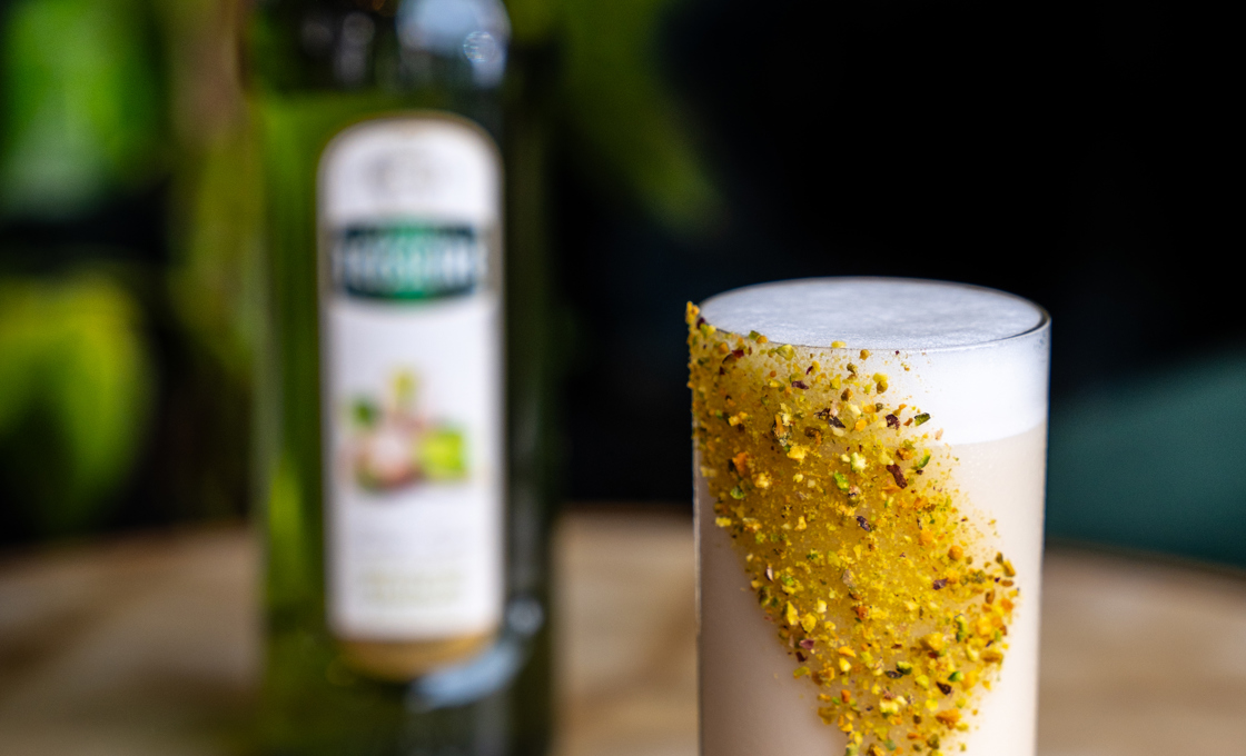 Atop a coffee table there is a tall thin highball glass, which contains a white drink and a is garnished with a pistachio stripe on the outside of the glass. In the background there is a bottle of Mathieu Teisseire pistachio syrup. 