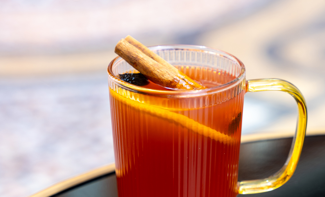 On the edge of a black coffee table there is a clear mug glass with a gold handle. The drink inside the glass is garnished with a cinnamon stick. 