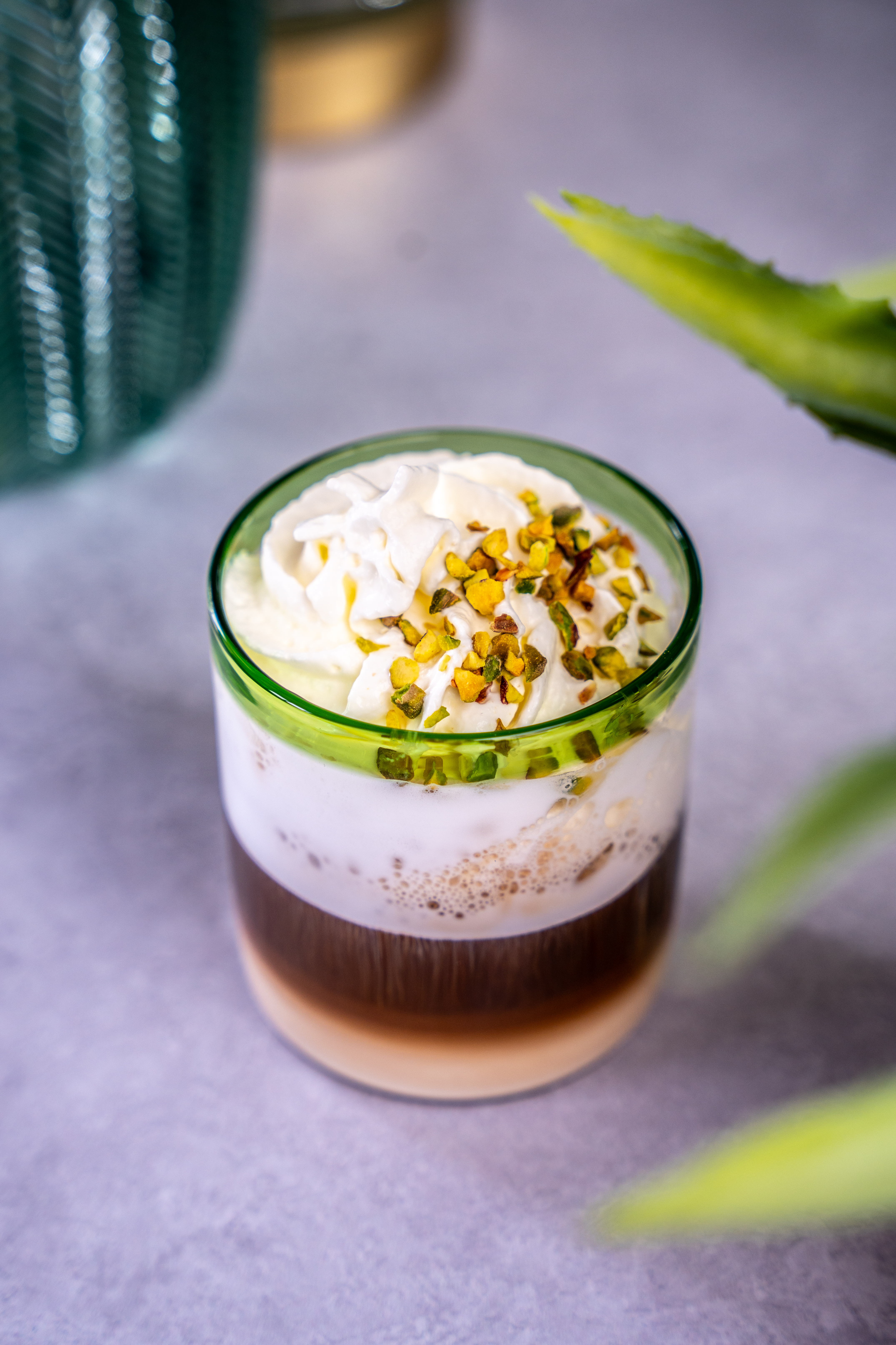 On top of a marble table there is a short tumbler glass that contains a coffee. The coffee is topped with whipped cream and crushed pistachio nuts. 