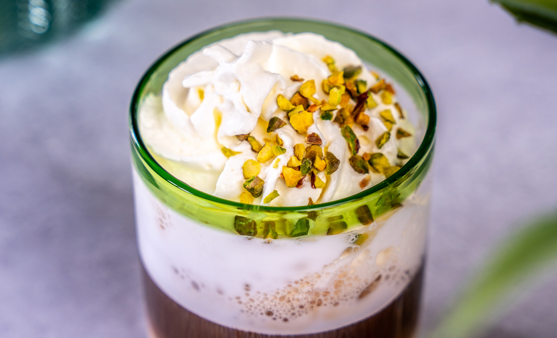 On top of a marble table there is a short tumbler glass that contains a coffee. The coffee is topped with whipped cream and crushed pistachio nuts. 