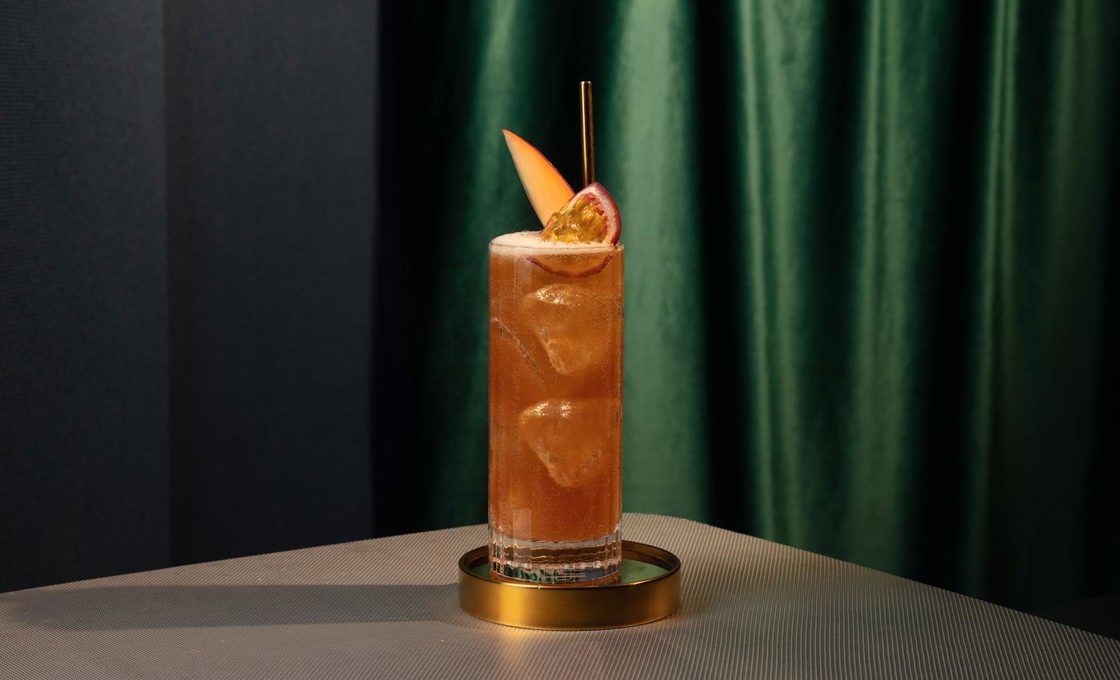 A highball glass is placed on a tall gold coaster. The glass contains a dark orange drink, ice, mango and pomegrate to garnish, and a gold metal straw.