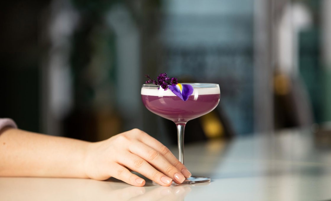 Inside a coupe glass there is a bright purple drink, garnished with purple flower leaves. A hand is reaching for the stem of the glass. In the background we can see an open window and some tall plants.