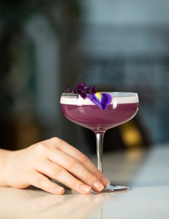 Inside a coupe glass there is a bright purple drink, garnished with purple flower leaves. A hand is reaching for the stem of the glass. In the background we can see an open window and some tall plants.