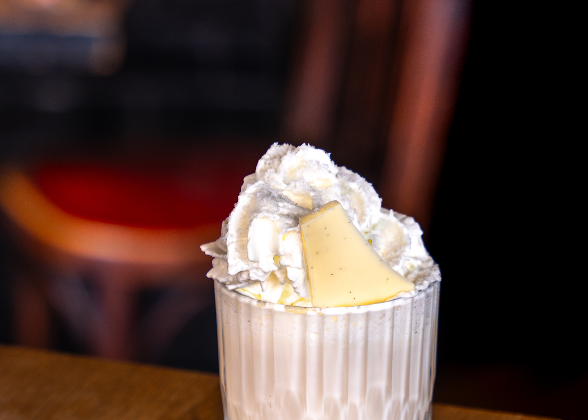 A short glass is placed on the edge of a wooden table. The glass is filled with a white drink, topped with whipped cream and white chocolate.