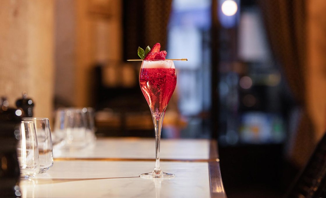 A champagne glass is filled with a sparkling red drink, and it garnished with a strawberry fan on a gold pick. In the background we see a bar and large doors.