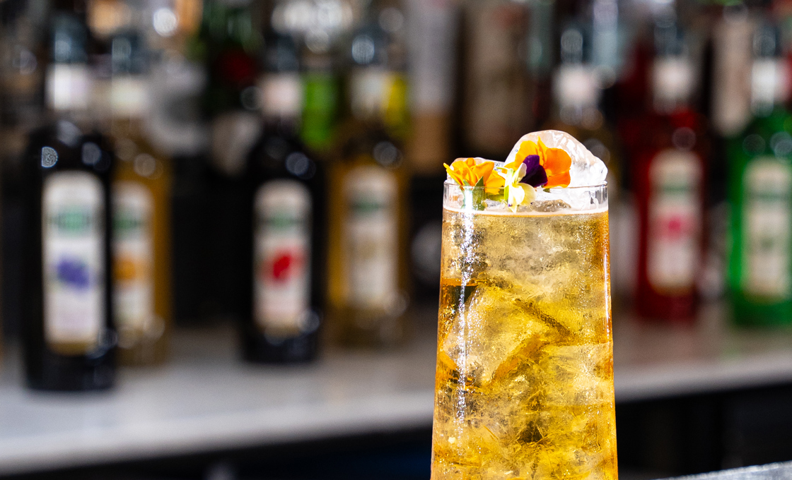 On top of a bar there is a tumbler glass filled with a pale yellow coloured drink. The drink is garnished with edible flowers. In the background of the bar we can see a range of Mathieu Teisseire bottles. 
