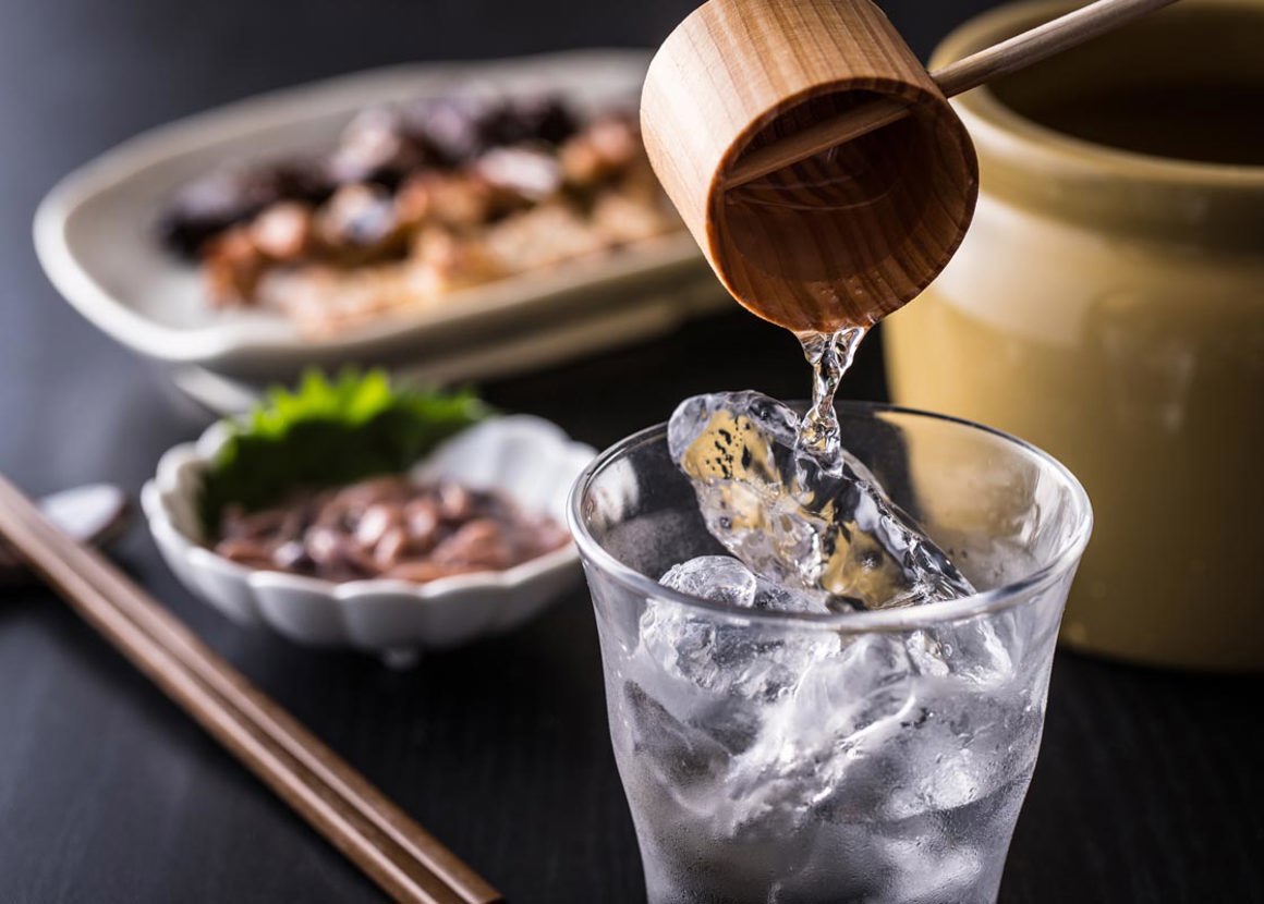 A clear liquid is being poured into a glass filled with ice. On the table in the background there are food dishes out of focus and wooden chopsticks.