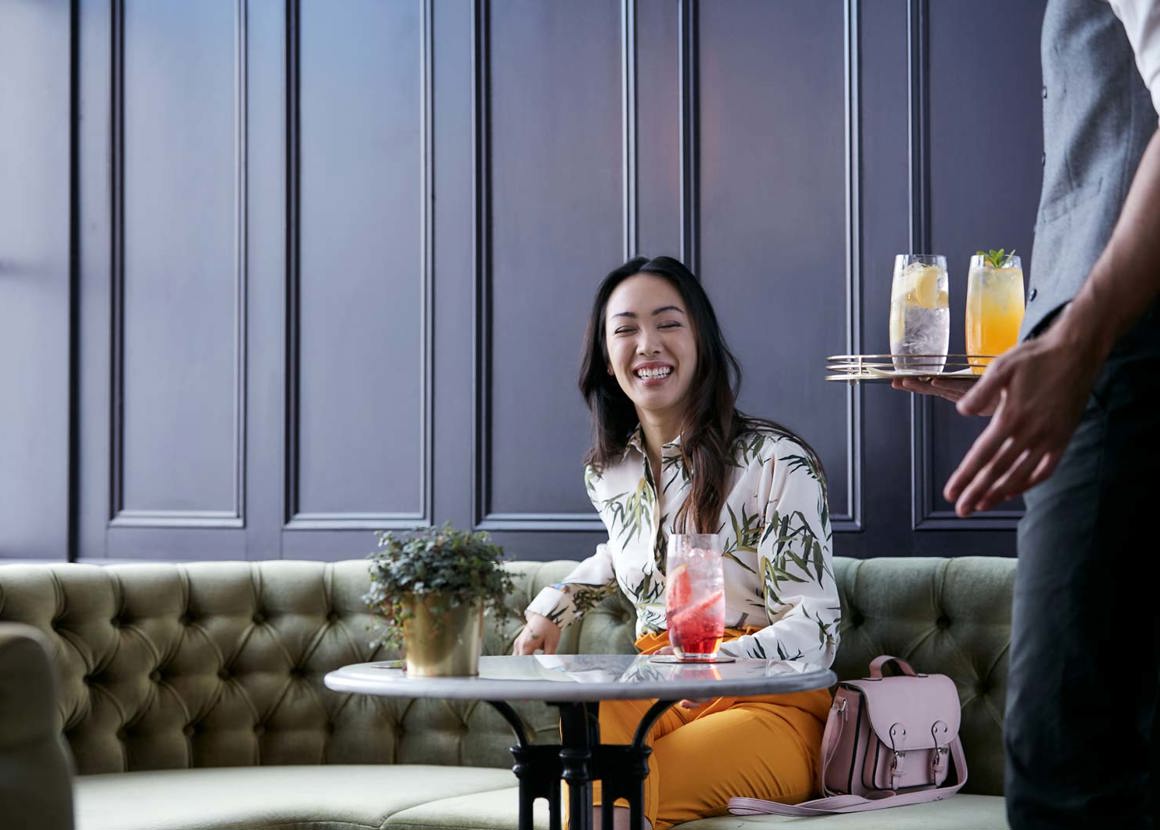 A woman sits smiling at a small marble table. A waiter in a waistcoat is bringing her drinks.